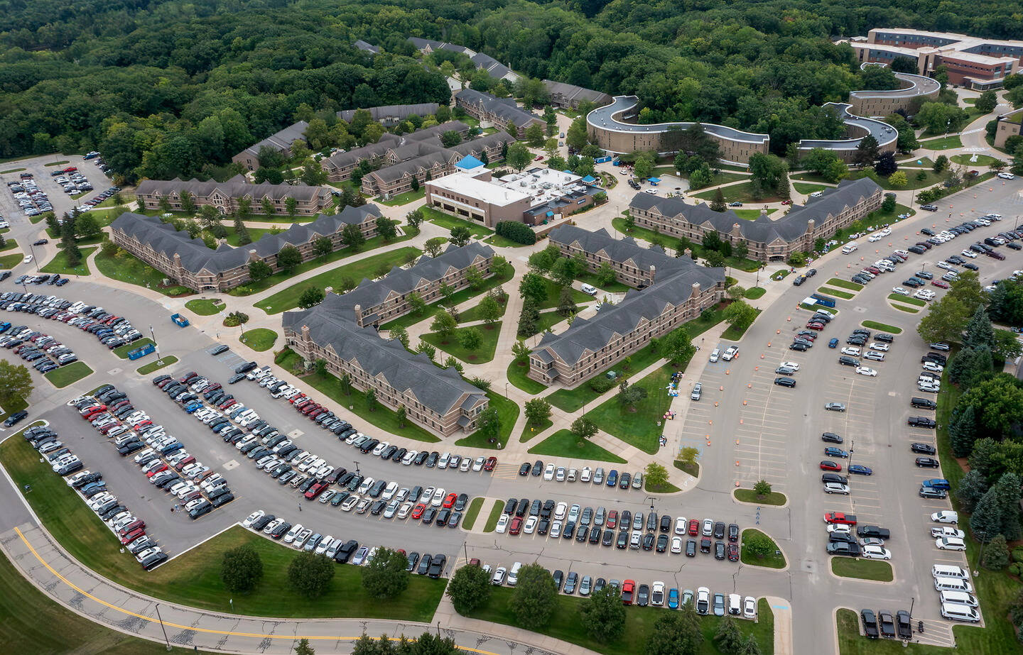 An aerial view of a large campus with multiple brick buildings, expansive parking lots filled with cars, and surrounding green trees.